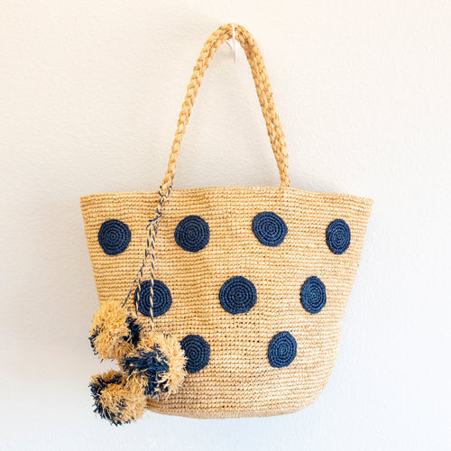 Blue polka-dot raffia tote hanging against a wall, showing braided handles and pom-poms 