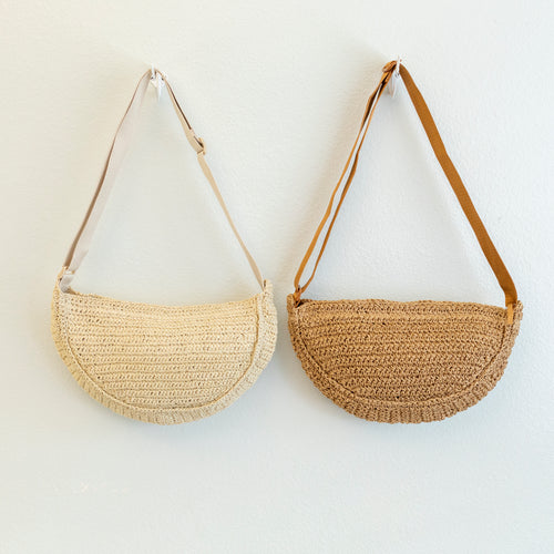 Cream and brown crescent woven straw crossbody bags hanging side by side against a white wall, each with a long adjustable strap 